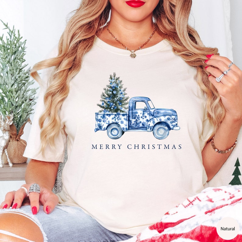 May include: White t-shirt with a blue and white floral print pickup truck carrying a blue and white floral print Christmas tree. The text "Merry Christmas" is printed below the truck.