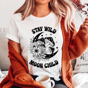 Puede incluir: Camiseta blanca con un dise&ntilde;o gr&aacute;fico negro que presenta una luna creciente con hongos y flores. El texto "Stay Wild Moon Child" est&aacute; impreso en negro.