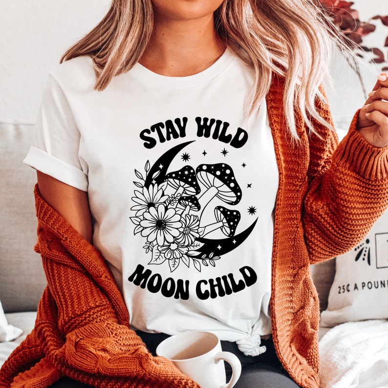 Puede incluir: Camiseta blanca con un dise&ntilde;o gr&aacute;fico negro que presenta una luna creciente con hongos y flores. El texto "Stay Wild Moon Child" est&aacute; impreso en negro.