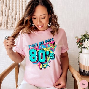 Pu&ograve; includere: Maglietta rosa con un design grafico che presenta il testo "Take Me Back to the 80's" in uno stile retr&ograve; con un pattino a rotelle e un nastro per cassette.