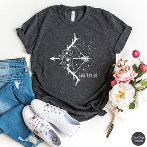May include: Dark grey heather t-shirt with a white floral design featuring a bow and arrow with the word "Sagittarius" underneath.