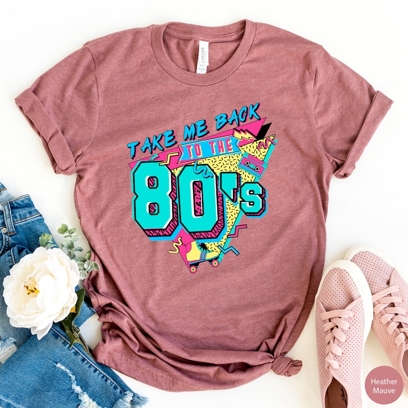 Pu&ograve; includere: Una t-shirt color malva melange con il testo "Take Me Back To The 80's" in un font retr&ograve; anni '80. La grafica include una musicassetta, pattini a rotelle e forme geometriche in rosa, blu e giallo.
