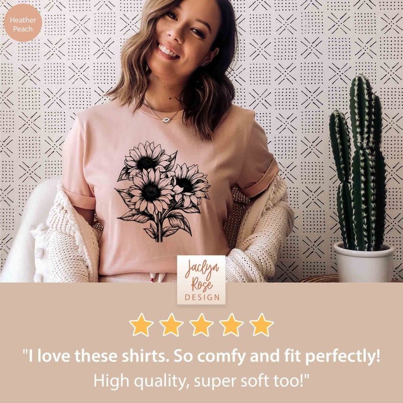 May include: Heather peach-colored t-shirt featuring a black and white sunflower graphic. The shirt has rolled-up sleeves and the text "Jaclyn Rose Design". The background has a white and black geometric pattern. The image also includes a cactus in a white pot.