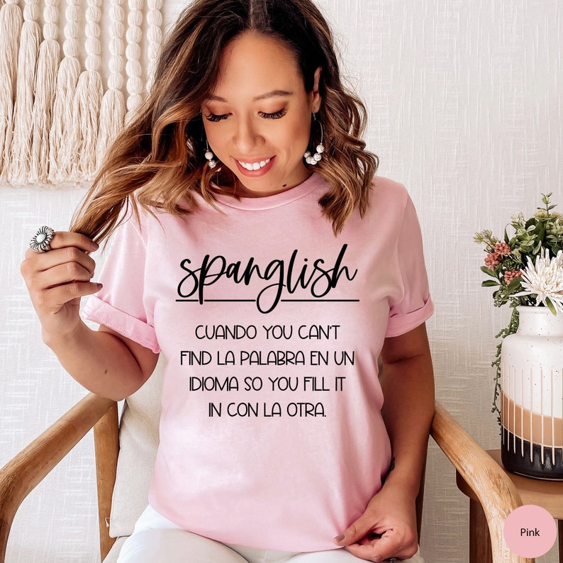 K&ouml;nnte beinhalten: Rosafarbenes T-Shirt mit schwarzem Text, der "Spanglish" lautet und eine Definition des Wortes in Spanisch und Englisch. Die Definition lautet: "CUANDO YOU CAN'T FIND LA PALABRA EN UN IDIOMA SO YOU FILL IT IN CON LA OTRA."