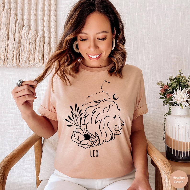 May include: A light pink t-shirt with a black graphic of a lion's head with a floral design and constellation. The text "LEO" is printed below the graphic.
