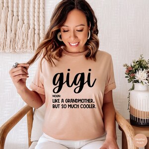 May include: A peach-coloured t-shirt with the word "gigi" written in black cursive lettering. The text "NOUN: LIKE A GRANDMOTHER, BUT SO MUCH COOLER." is written in black below the word "gigi".