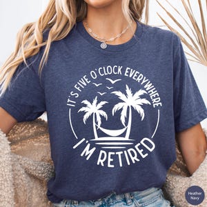 May include: Heather navy blue t-shirt with a white graphic. The graphic features two palm trees, a hammock, and the text "It's Five O'Clock Everywhere, I'm Retired". The shirt is a crew neck.