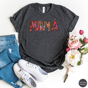 May include: Dark grey heather t-shirt with the word "MOMA" in colorful letters. The letters are decorated with floral and religious imagery. The shirt is displayed on a white background with a pair of white sneakers and a bouquet of pink flowers.