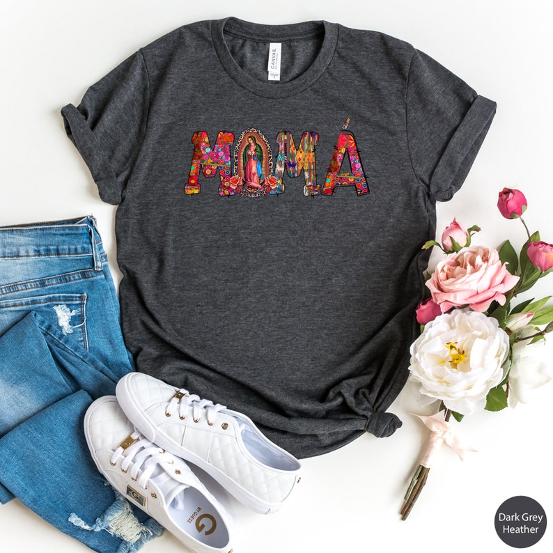 May include: Dark grey heather t-shirt with the word "MOMA" in colorful letters. The letters are decorated with floral and religious imagery. The shirt is displayed on a white background with a pair of white sneakers and a bouquet of pink flowers.