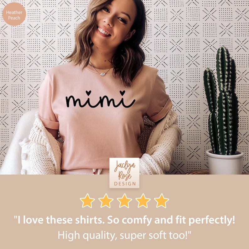 May include: A pink t-shirt with the word "mimi" written in black cursive with two small hearts above the word. The shirt is being worn by a person and has a white background with a black and white pattern.