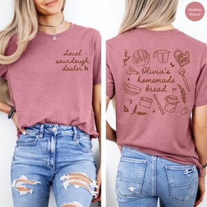 Pu&ograve; includere: T-shirt color malva melange con la scritta "Local sourdough dealer" sul davanti. Il retro della maglietta presenta un'illustrazione marrone di strumenti per la panificazione e la scritta "Olivia's homemade bread".