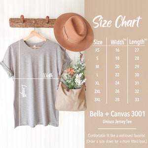 May include: Size chart for a Bella + Canvas 3001 Unisex Jersey Tee. The chart shows the width and length in inches for sizes XS to 3XL. The image also shows a gray t-shirt hanging on a wooden rack.