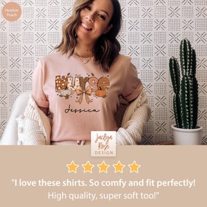 May include: A pink t-shirt with a fall-themed "Nurse" graphic featuring pumpkins, leaves, and a bow. The text "Jessica" is printed below the graphic. The shirt is on a model and has a five-star rating with the text "I love these shirts. So comfy and fit perfectly! High quality, super soft too!"