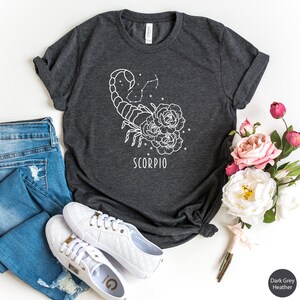 Pu&ograve; includere: T-shirt grigio scuro melange con un contorno bianco di uno scorpione e rose con la parola "SCORPIO" stampata sotto.