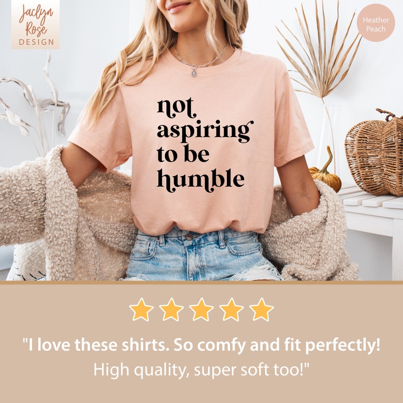 May include: A light pink t-shirt with the text "not aspiring to be humble" printed in black. The shirt is being modeled by a person wearing blue jeans. The text "Heather Peach" is visible in the upper right corner of the image.