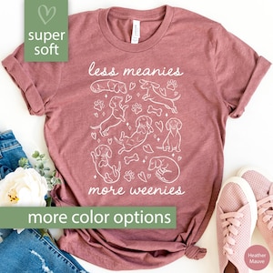 Op de afbeelding: Heather mauve t-shirt met de tekst "less meanies more weenies" en een witte afbeelding van teckels, pootafdrukken en botten. Het shirt is gemaakt van superzacht materiaal.
