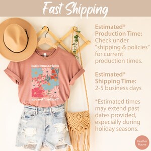 May include: A heather mauve t-shirt with a floral design and the text "basic human rights are not 'radical'" printed on the front. The shirt is displayed on a wooden hanger with a straw hat and a woven bag.