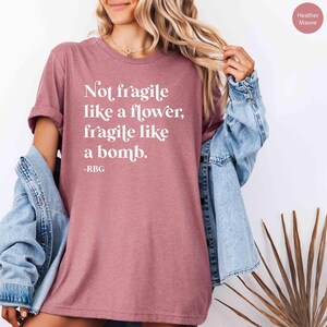 May include: A heather mauve t-shirt with the text "Not fragile like a flower, fragile like a bomb. -RBG" printed in white.