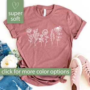 May include: A mauve t-shirt with a white outline of a row of flowers. The text "super soft" is in a green heart shape in the top left corner. The text "click for more color options" is in a green rectangle at the bottom of the image.