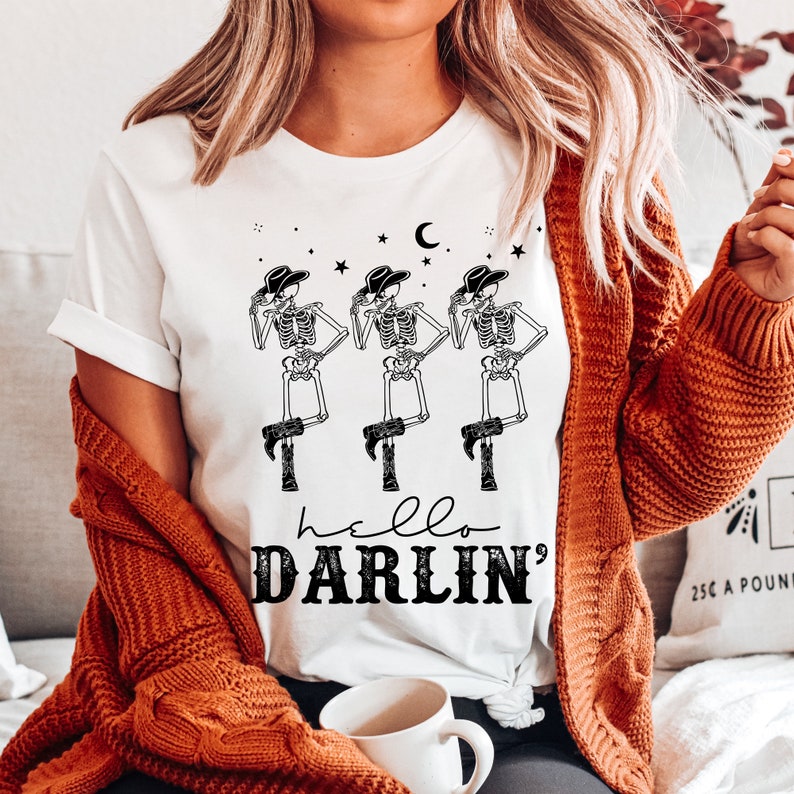 May include: White t-shirt with a graphic of three skeletons wearing cowboy hats and boots. The text "hello darlin'" is printed below the skeletons.