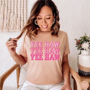 May include: A light pink t-shirt with the text "YEE HAW" repeated three times in a pink gradient. The text is stylized with small white hearts.