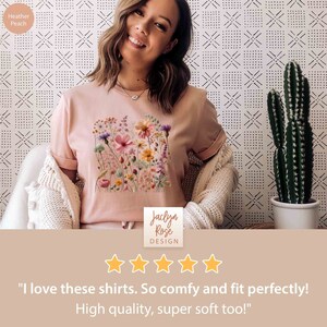 K&ouml;nnte beinhalten: Eine Frau tr&auml;gt ein T-Shirt in der Farbe Heidelbeer-Peach mit einem Blumenmuster. Das Shirt hat einen Rundhalsausschnitt und kurze &Auml;rmel. Der Blumendruck ist in den Farben Rosa, Gelb und Gr&uuml;n gehalten. Das Shirt stammt von Jaclyn Rose Design. Der Text "Ich liebe diese Shirts. So bequem und perfekt sitzend! Hochwertige Qualit&auml;t, super weich auch!" befindet sich unter dem Bild.