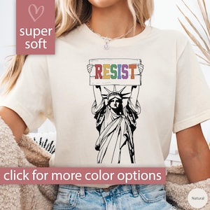 May include: A white t-shirt with a black and white illustration of the Statue of Liberty holding a sign that says "RESIST" in rainbow colors.