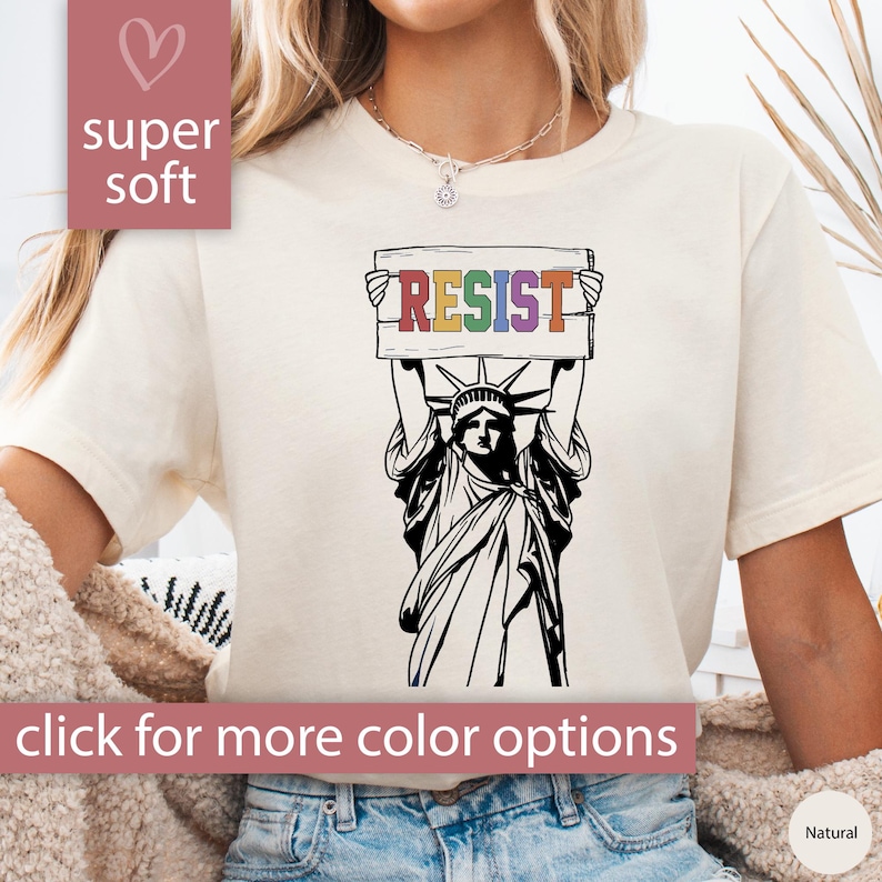 May include: A white t-shirt with a black and white illustration of the Statue of Liberty holding a sign that says "RESIST" in rainbow colors.