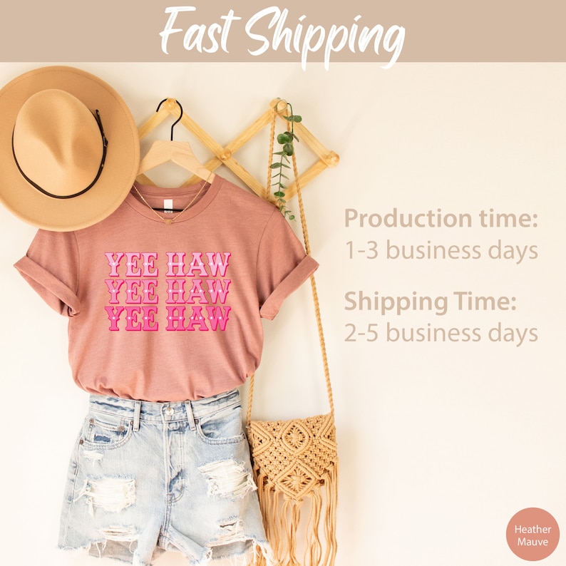 May include: A heather mauve t-shirt with the text "YEE HAW" repeated three times in pink. The shirt is hanging on a wooden hanger with a straw hat and a woven bag.