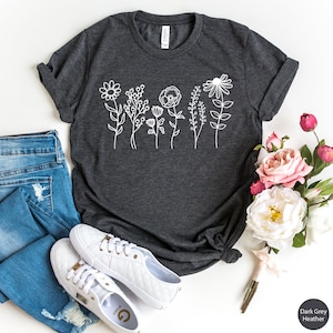 May include: A dark grey heather t-shirt with a white graphic of stylized flowers. The shirt is laid flat on a white surface with a pair of white sneakers and a bouquet of pink flowers.