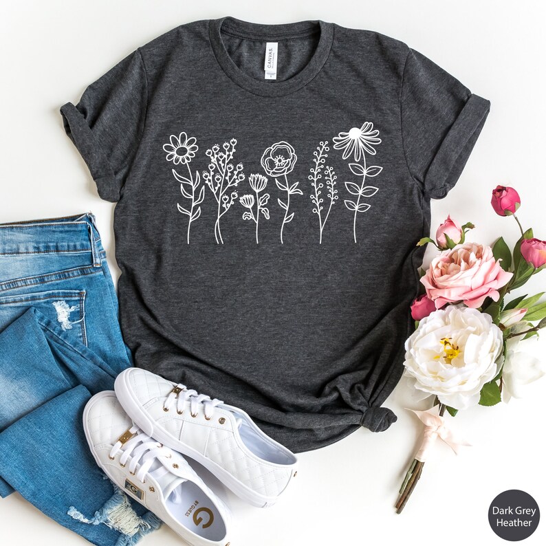 May include: A dark grey heather t-shirt with a white graphic of stylized flowers. The shirt is laid flat on a white surface with a pair of white sneakers and a bouquet of pink flowers.