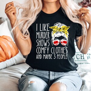 May include: Black t-shirt with white text that reads "I like murder shows, comfy clothes and maybe 3 people". A cartoon woman with a messy bun and sunglasses is wearing a yellow "Do Not Cross" tape headband.