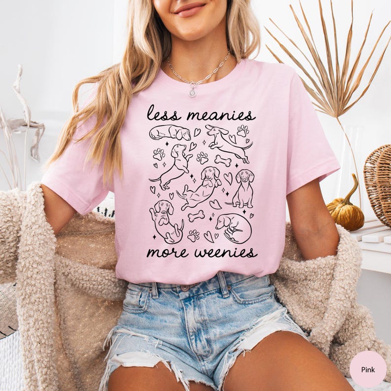 Op de afbeelding: Lichtroze T-shirt met de tekst "less meanies more weenies" boven en onder een zwarte illustratie van teckels, pootafdrukken en botten. Het shirt heeft korte mouwen.