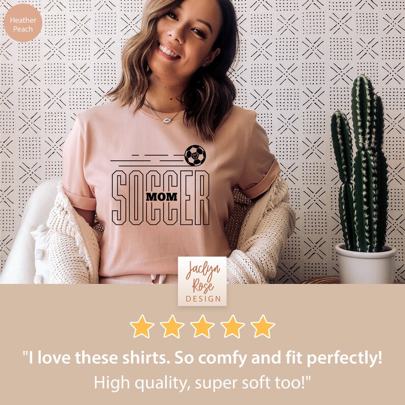 Pu&ograve; includere: Una maglietta rosa con una grafica nera che dice "Soccer Mom" con un pallone da calcio. La maglietta &egrave; indossata da una persona. Il testo "Jaclyn Rose Design" &egrave; anche visibile nell'immagine. L'immagine ha una valutazione a 5 stelle.