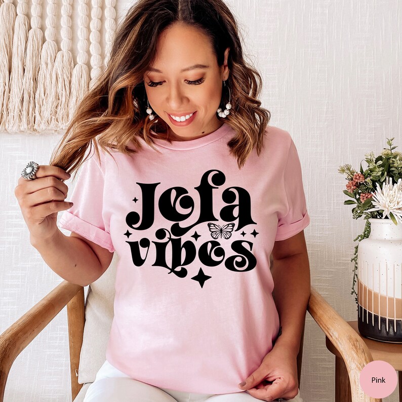 May include: A light pink t-shirt with the words "Jefa Vibes" in a black, retro font, with a butterfly graphic. The shirt is short-sleeved and has a relaxed fit. The model is wearing the shirt.