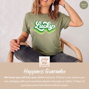 May include: A green t-shirt with the word "Lucky" in a retro, multi-colored font. The shirt is being modeled by a person sitting on a wooden chair. The text "Heather Olive" is in the top right corner of the image. The text "Jaclyn Rose DESIGN" is in the bottom center of the image. The text "Happiness Guarantee" is in the bottom center of the image. The text "We know you will love your shirt! However, if there is any reason you are unhappy with your purchase please message us within 14 days of purchase and a full refund will be issued." is in the bottom center of the image.