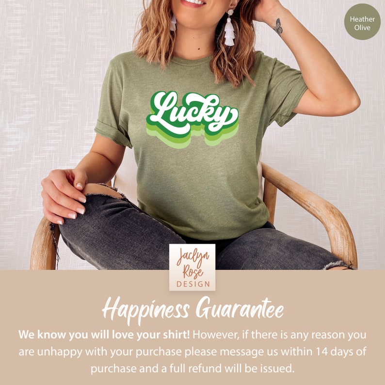 May include: A green t-shirt with the word "Lucky" in a retro, multi-colored font. The shirt is being modeled by a person sitting on a wooden chair. The text "Heather Olive" is in the top right corner of the image. The text "Jaclyn Rose DESIGN" is in the bottom center of the image. The text "Happiness Guarantee" is in the bottom center of the image. The text "We know you will love your shirt! However, if there is any reason you are unhappy with your purchase please message us within 14 days of purchase and a full refund will be issued." is in the bottom center of the image.