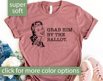 Puede incluir: Una camiseta de color malva con una ilustración vintage de una mujer y el texto "Grab Him By The Ballot."