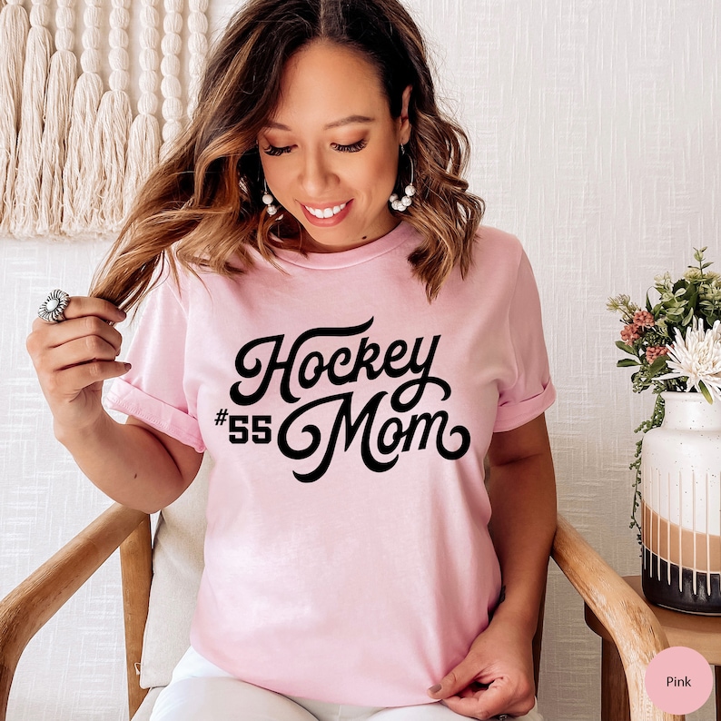 Peut inclure: T-shirt rose avec un texte noir qui dit "Hockey Mom #55" dans une police r&eacute;tro.