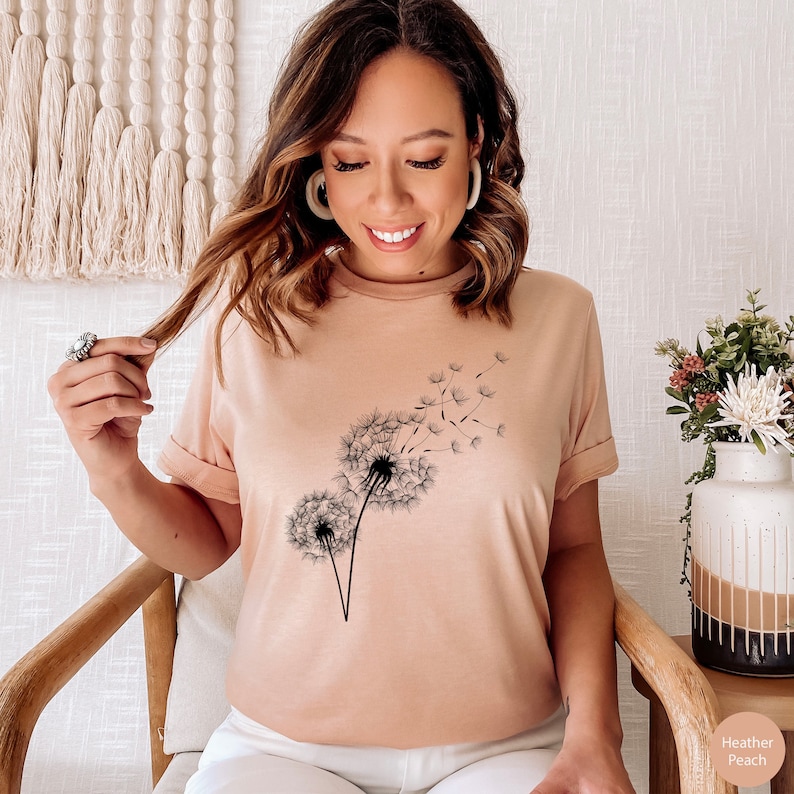 May include: A heather peach colored t-shirt with a black dandelion graphic. The dandelion has seeds blowing in the wind.