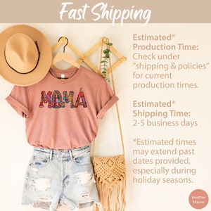 May include: A heather mauve t-shirt with the word "MAMA" spelled out in colorful mosaic letters. The letters are decorated with images of religious figures. The shirt is hanging on a wooden hanger with a straw hat and a woven bag.