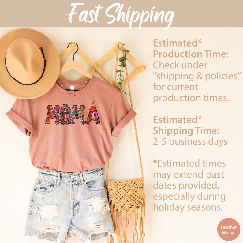 May include: A heather mauve t-shirt with the word "MAMA" spelled out in colorful mosaic letters. The letters are decorated with images of religious figures. The shirt is hanging on a wooden hanger with a straw hat and a woven bag.