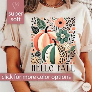 May include: A beige t-shirt with a graphic print of pumpkins, flowers, and leaves in shades of green, orange, and brown. The text "HELLO FALL" is printed below the graphic. The shirt is labeled "super soft" and "Natural".