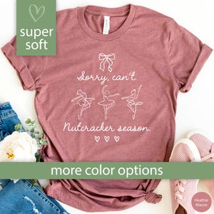 May include: Heather mauve t-shirt with white text that reads "Sorry, can't. Nutcracker season." and features a bow and three ballerina figures. The shirt has rolled sleeves and is labeled "super soft."