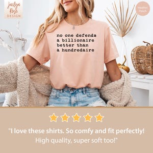 K&ouml;nnte beinhalten: Ein pinkfarbenes T-Shirt mit dem Aufdruck "no one defends a billionaire better than a hundredaire". Das Shirt wird von einer Person getragen und vor dem Hintergrund einer wei&szlig;en Wand und einem Weidenkorb mit Herbstdekoration fotografiert.