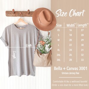 May include: Size chart for a Bella + Canvas 3001 Unisex Jersey Tee. The chart shows the width and length in inches for sizes XS to 3XL. The image also shows a gray t-shirt hanging on a wooden rack.