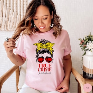 May include: Pink t-shirt with a graphic of a woman with a messy bun wearing sunglasses and a yellow "Do Not Cross" crime scene tape headband. The text "True Crime Junkie" is printed below the graphic.