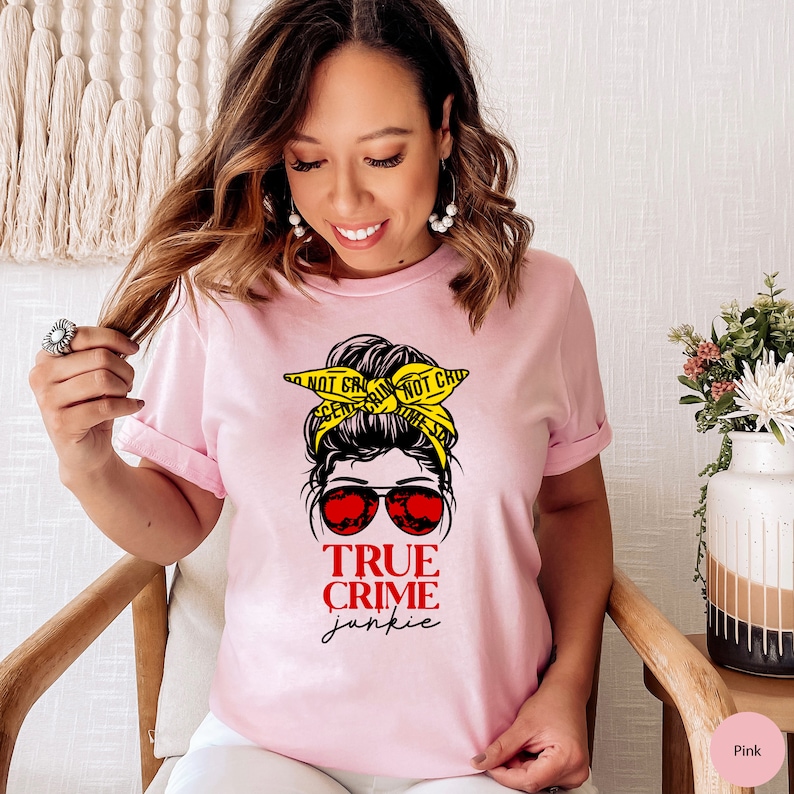 May include: Pink t-shirt with a graphic of a woman with a messy bun wearing sunglasses and a yellow "Do Not Cross" crime scene tape headband. The text "True Crime Junkie" is printed below the graphic.