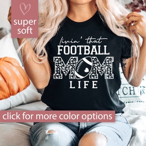 May include: Black t-shirt with white text that reads "livin' that FOOTBALL MOM LIFE" with a football and leopard print design.