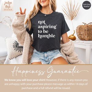 May include: A dark grey heather t-shirt with the text "not aspiring to be humble" printed in white. The shirt is being worn by a person with long blonde hair. The text "Happiness Guarantee" is printed in a cursive font on a light brown background.